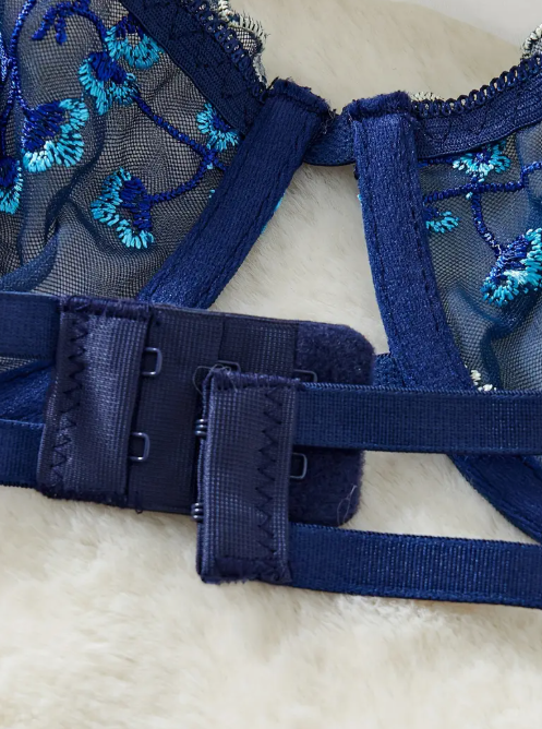 A navy blue sheer mesh lingerie set from The Seventh Muse with blue and white floral embroidery, including a plunge bra with cut-outs and an adjustable thong.