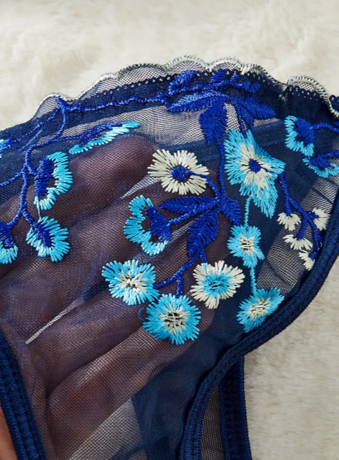 A navy blue sheer mesh lingerie set from The Seventh Muse with blue and white floral embroidery, including a plunge bra with cut-outs and an adjustable thong.
