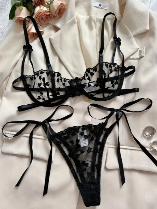 A black sheer mesh bra and matching thong set from The Seventh Muse with floral/heart embroidery and decorative satin ribbon ties, displayed on a cream blazer.
