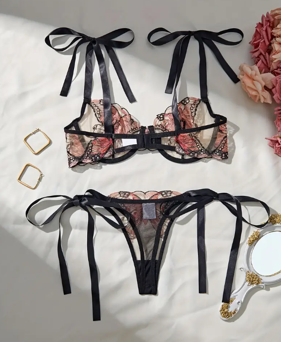 A lingerie set with butterfly embroidery, consisting of a sheer black bralette with bow ties on the shoulders and a matching thong, displayed on a white background with accessories in the background.