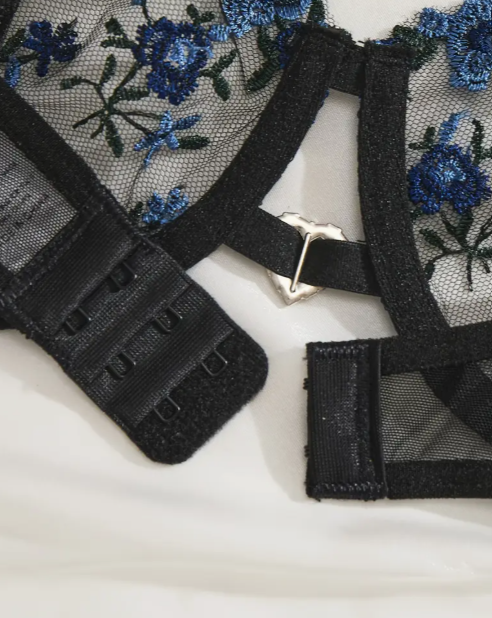 Close-up of black mesh bra with floral blue embroidery and heart-shaped metallic charm – hook-and-eye clasp detail by The Seventh Muse