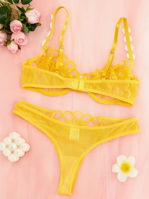 A yellow lingerie set with floral embroidery, displayed on a pink background with decorative flowers and petals around it.