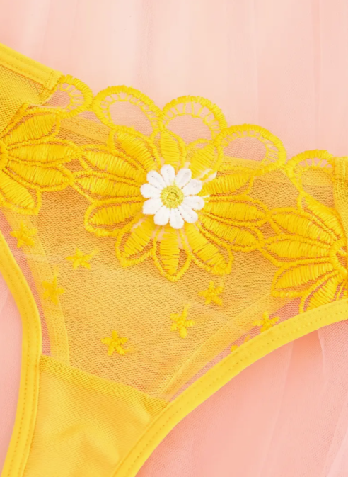 A yellow lingerie set with floral embroidery, displayed on a pink background with decorative flowers and petals around it.