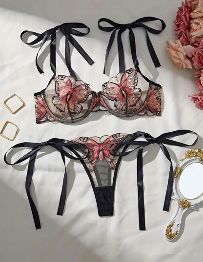 A lingerie set with butterfly embroidery, consisting of a sheer black bralette with bow ties on the shoulders and a matching thong, displayed on a white background with accessories in the background.
