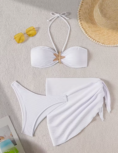 White three-piece bikini set for women featuring a starfish embellishment, halter neck bandeau top, matching bikini bottom, and wrap cover-up skirt – chic beachwear from The Seventh Muse.